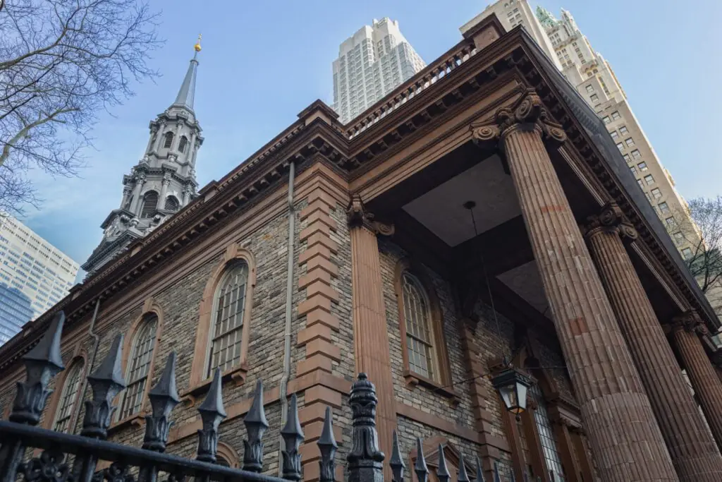 Low-angle view of St. Paul’s Chapel with its stone facade, classical columns, steeple, and surrounding Manhattan skyscrapers.